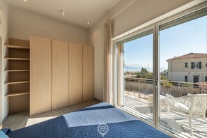 A bedroom with a large window, a bed, a wardrobe, and a balcony with a view of the sea.