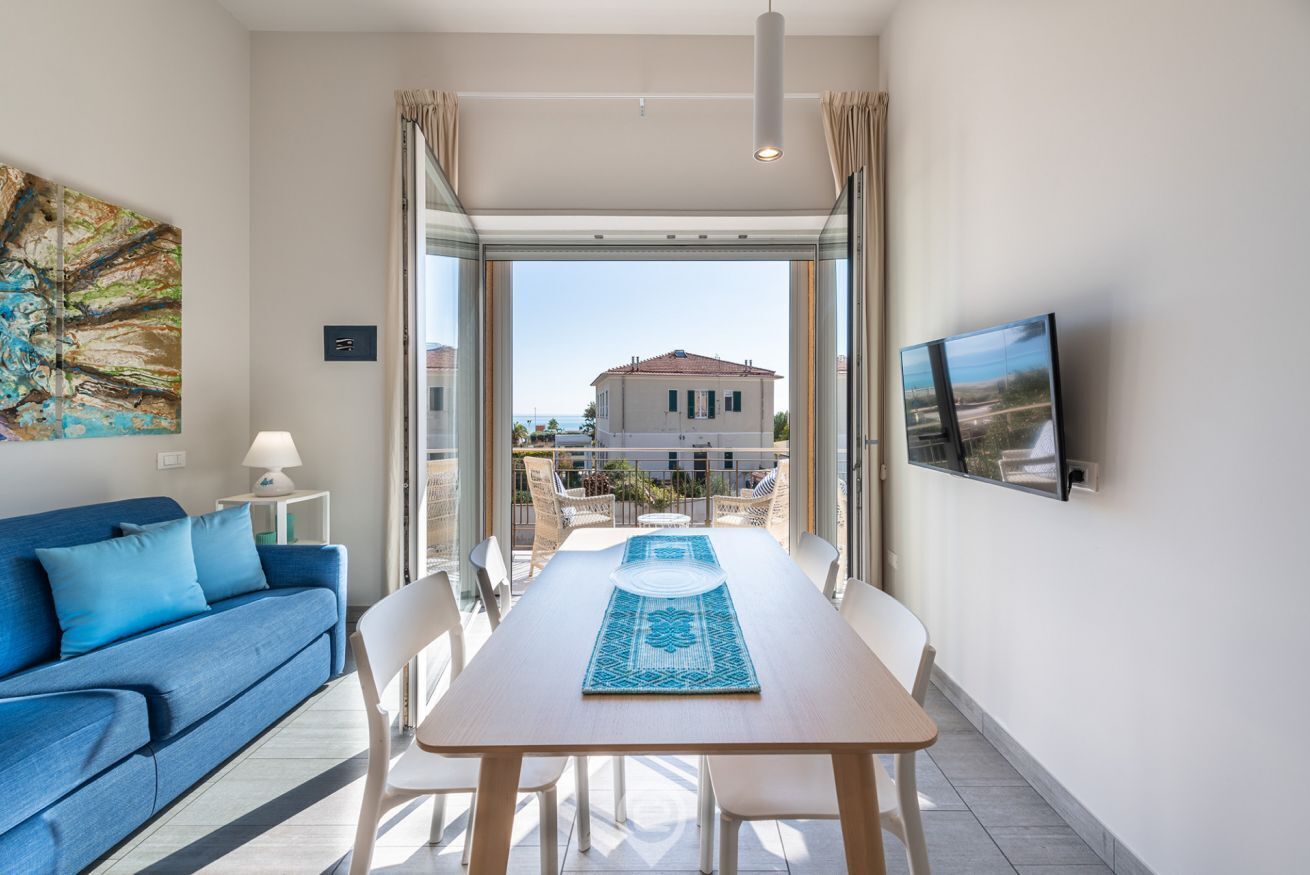 Un soggiorno moderno con un divano blu, un tavolo da pranzo in legno con sedie, una TV a parete e vista sul balcone esterno.