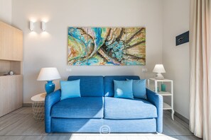 A modern living room with a blue sofa, two side tables with lamps, a large abstract painting on the wall, and a wooden cabinet. - Poetto Sea Breeze - Blu (Cagliari)