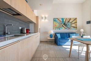 A modern kitchen with a built-in stove, a sink, and a coffee machine. There is a blue sofa and a small round table with a lamp.