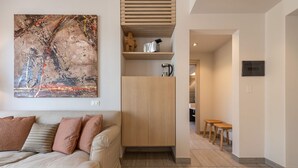 Een moderne woonkamer met een beige bank, een groot abstract schilderij, een houten kast, en een keuken op de achtergrond.