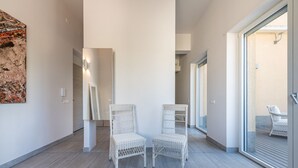 Een ruime hal met twee witte rieten stoelen, een groot abstract schilderij aan de linkerkant, en een glazen schuifdeur die leidt naar een buitenruimte met een stoel.