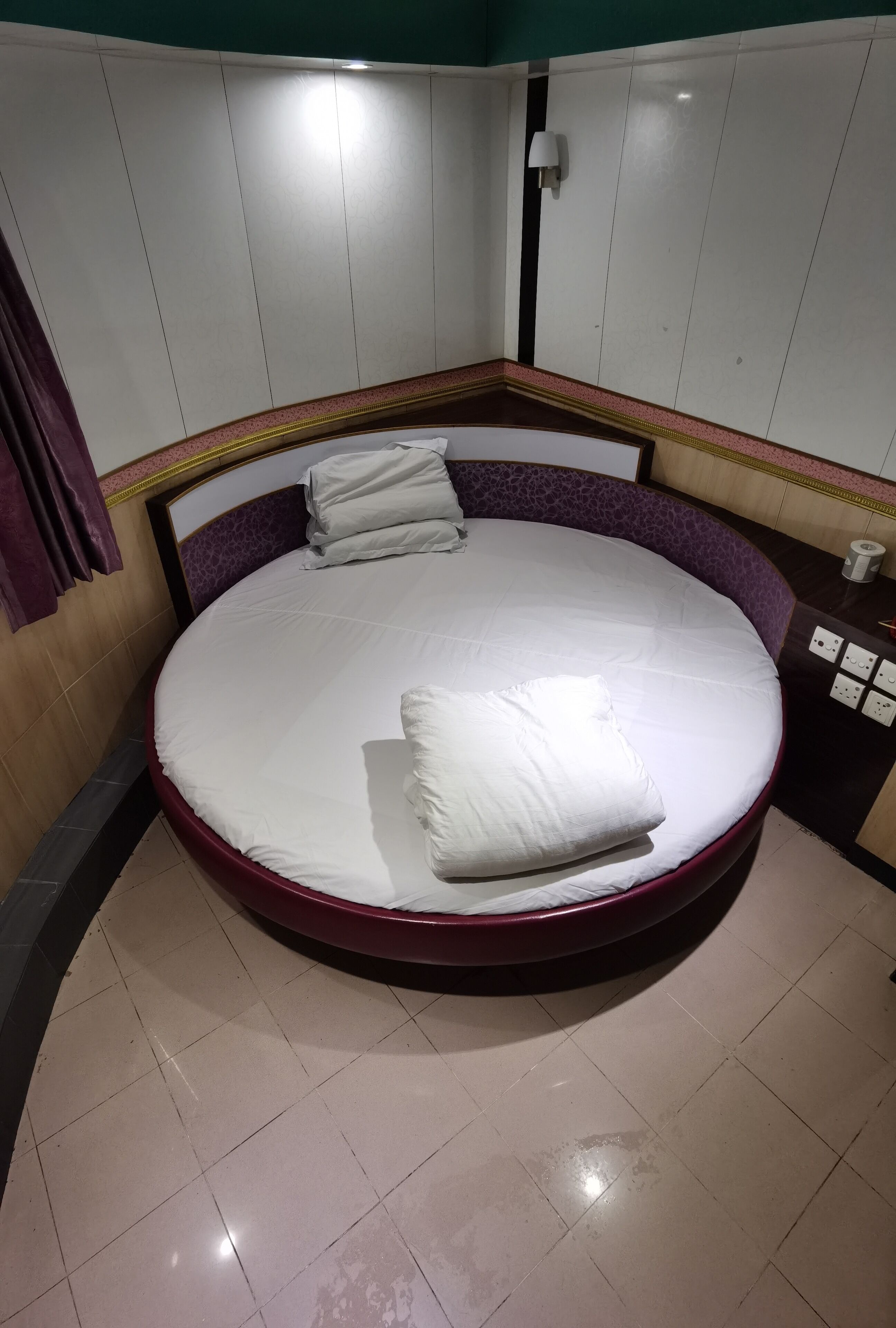 A round bed with a wooden frame and a purple headboard, white bedding, and a small bedside table with a lamp and a tissue box.