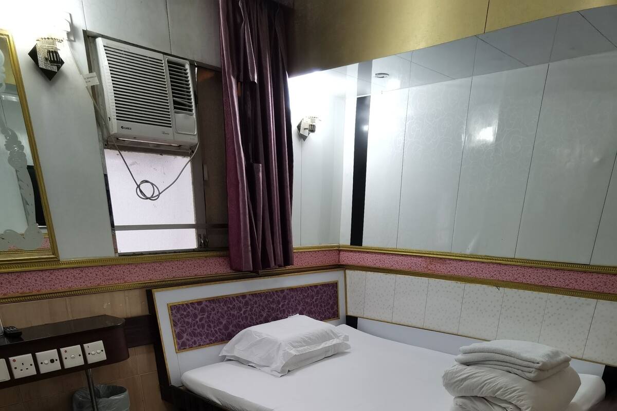A hotel room with a bed, a small stool, a trash bin, and an air conditioning unit.
