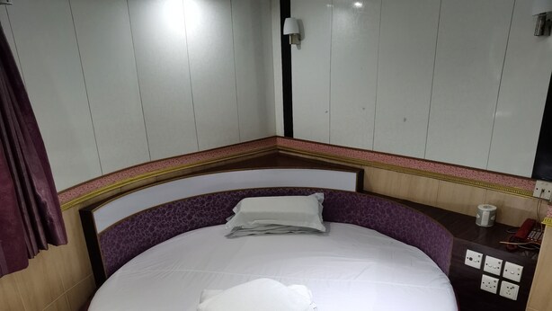 A round bed with white bedding and pillows, a headboard with a purple and yellow pattern, and a wall-mounted light fixture. - spanish hostel (Kowloon)