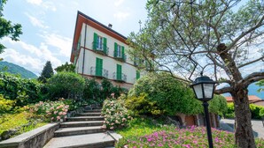 A building with a staircase leading to a balcony, surrounded by flowering plants and a tree. - Green House Regatola by Wonderful Italy (Bellagio)