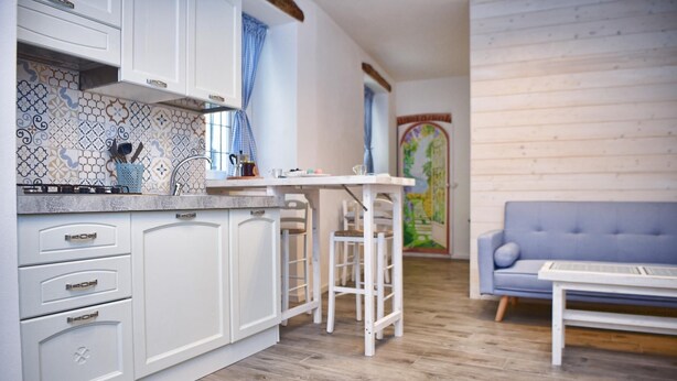 A modern kitchen with white cabinets, a tiled backsplash, and a bar stool. There is a blue sofa and a small table in the living area. - Ca de Sota by Wonderful Italy (Bellagio)