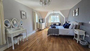 Un dormitorio con piso de madera, una cama con ropa de cama azul y blanca, una tocador blanco con espejos, una araña y una ventana con cortinas.