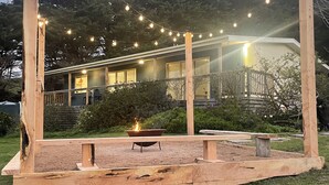 Une terrasse en bois avec un foyer, entourée d’une pelouse et illuminée de guirlandes lumineuses.