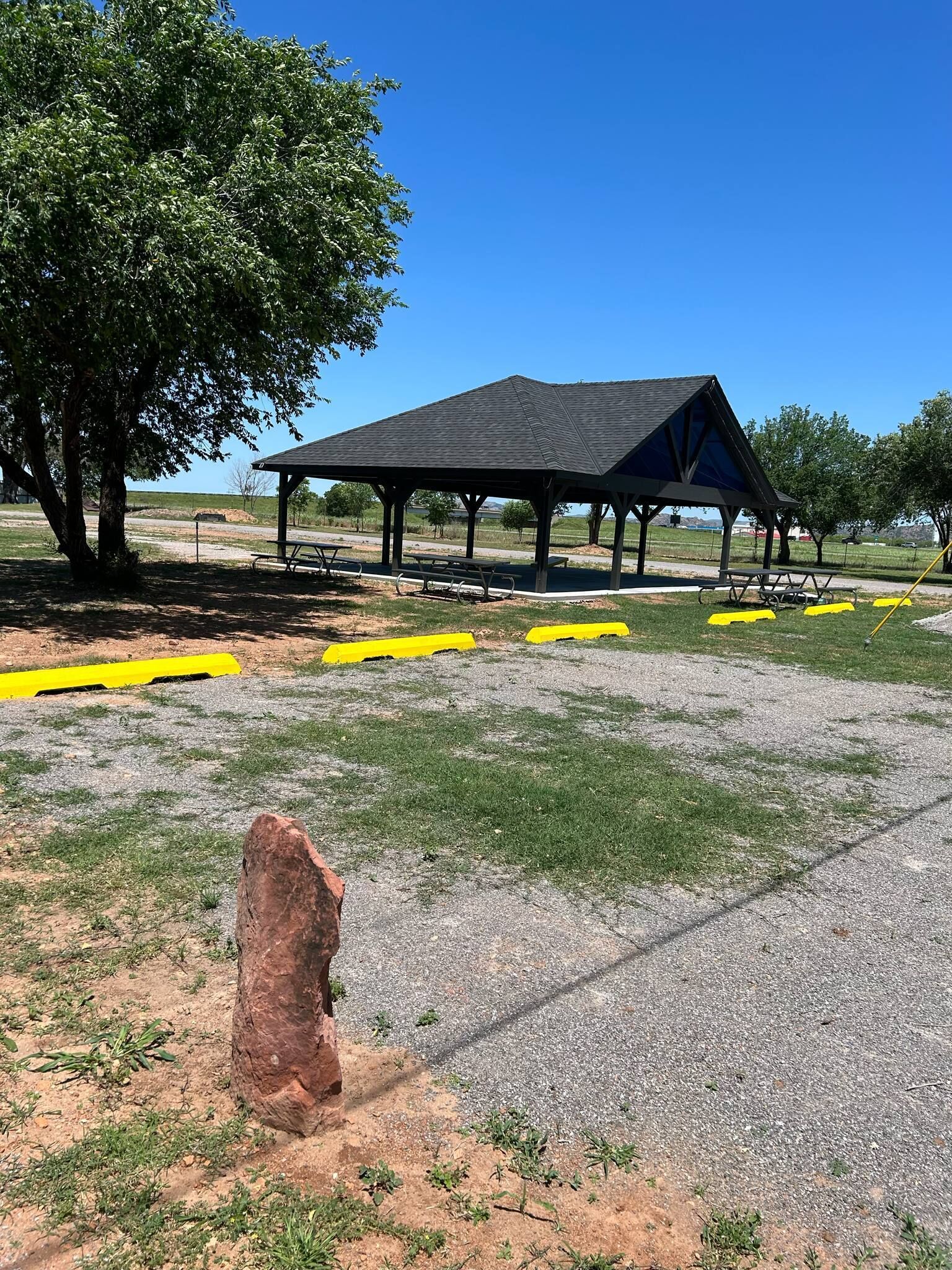 A covered pavilion with picnic tables, a large tree, and yellow safety markings on the ground.