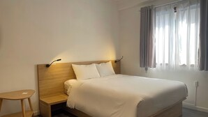 Een netjes opgemaakt bed met een houten hoofdbord en nachtkastjes, een klein houten krukje en een raam met gordijnen.