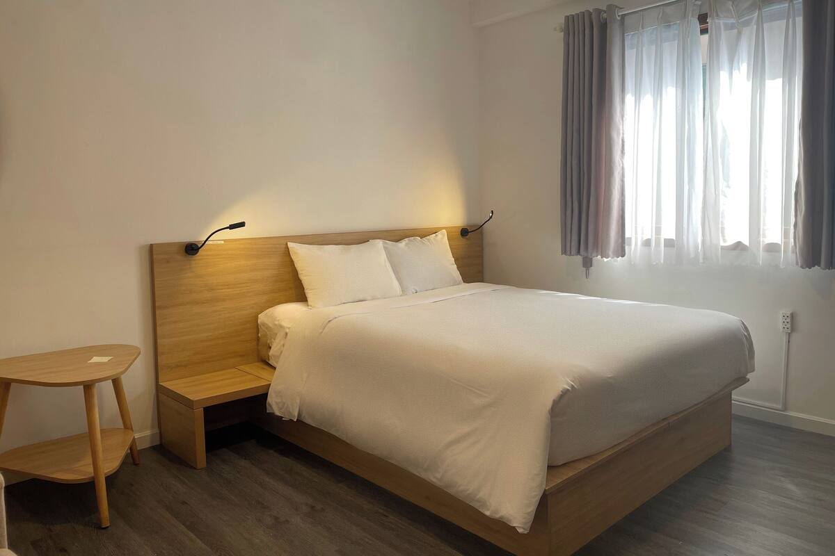 A neatly made bed with a wooden headboard and bedside tables, a small wooden stool, and a window with curtains.