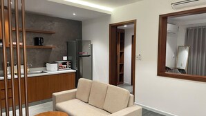 Een moderne woonkamer met een beige bank, een houten eettafel en een keuken met een koelkast en koffiezetapparaat.