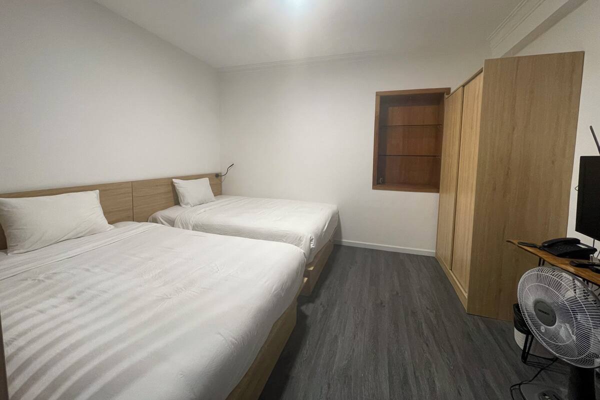 A room with two beds, a wooden wardrobe, a desk with a computer and fan, and a small window.