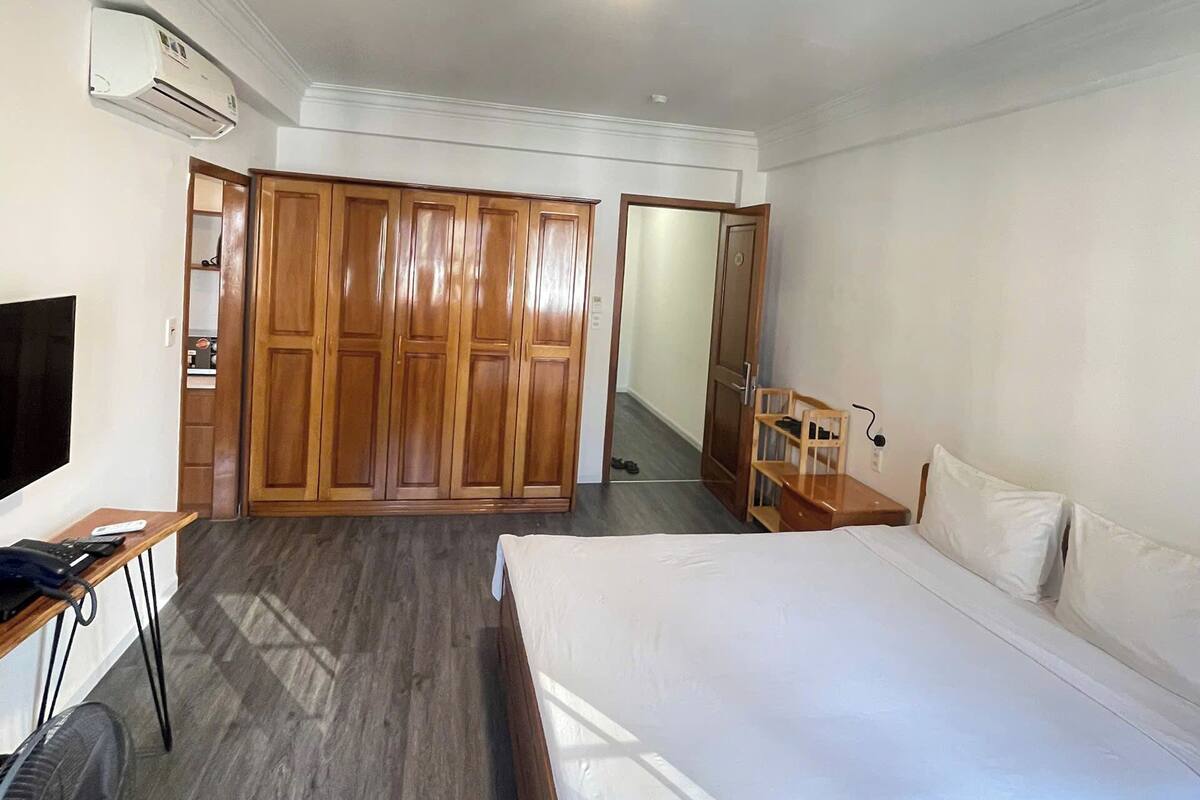 A hotel room with a bed, a desk with a chair, a wardrobe, and an air conditioning unit.