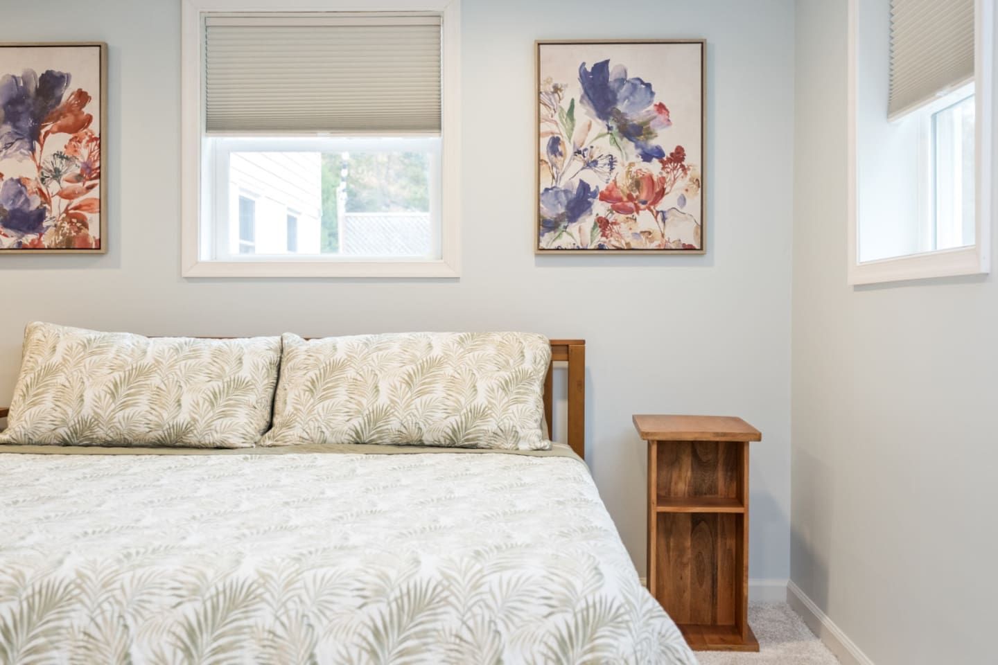 Quarto com uma cama, duas almofadas, uma mesa de cabeceira e duas obras de arte com flores emolduradas na parede.