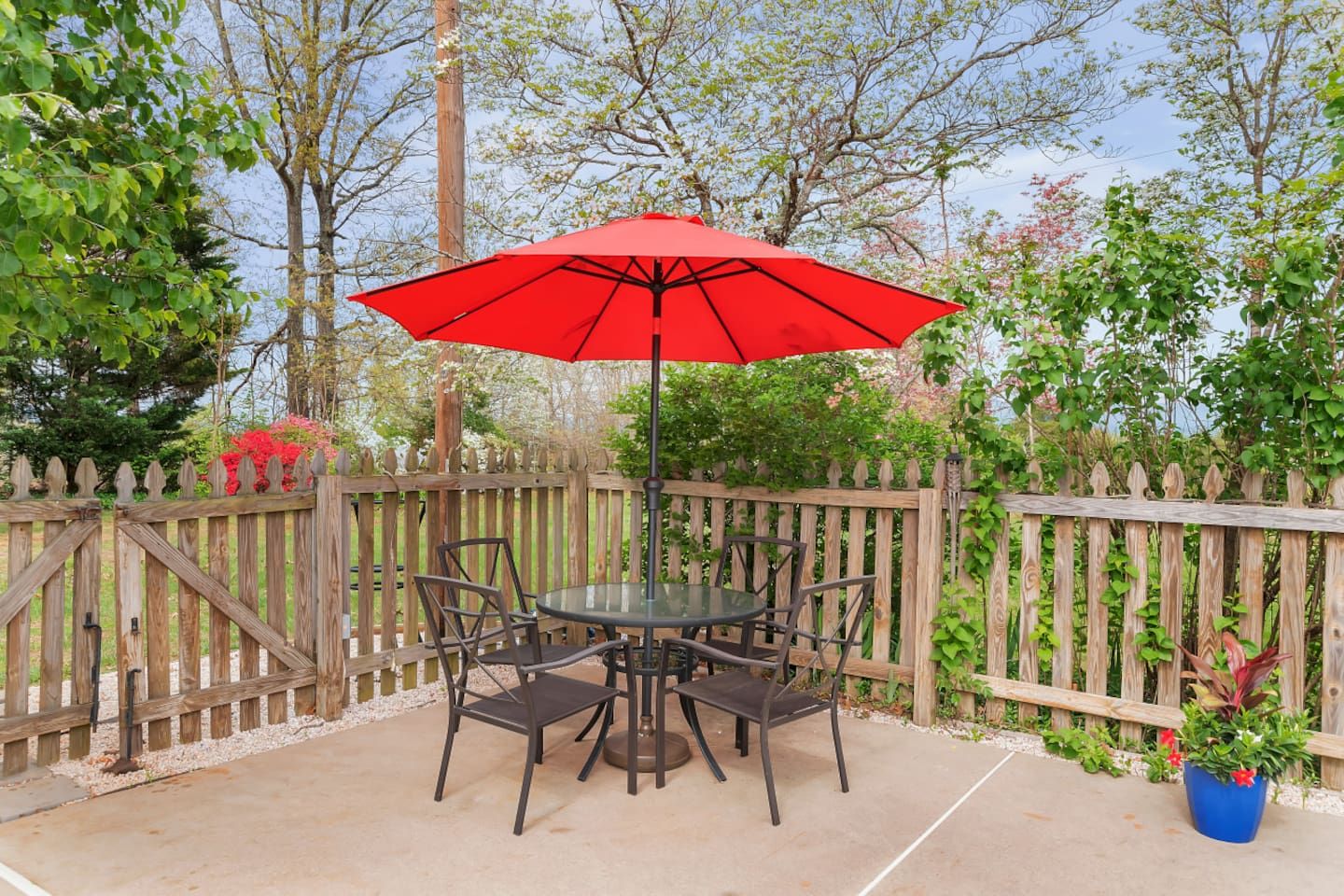 A patio with a round table and chairs, a red umbrella, and a wooden fence.