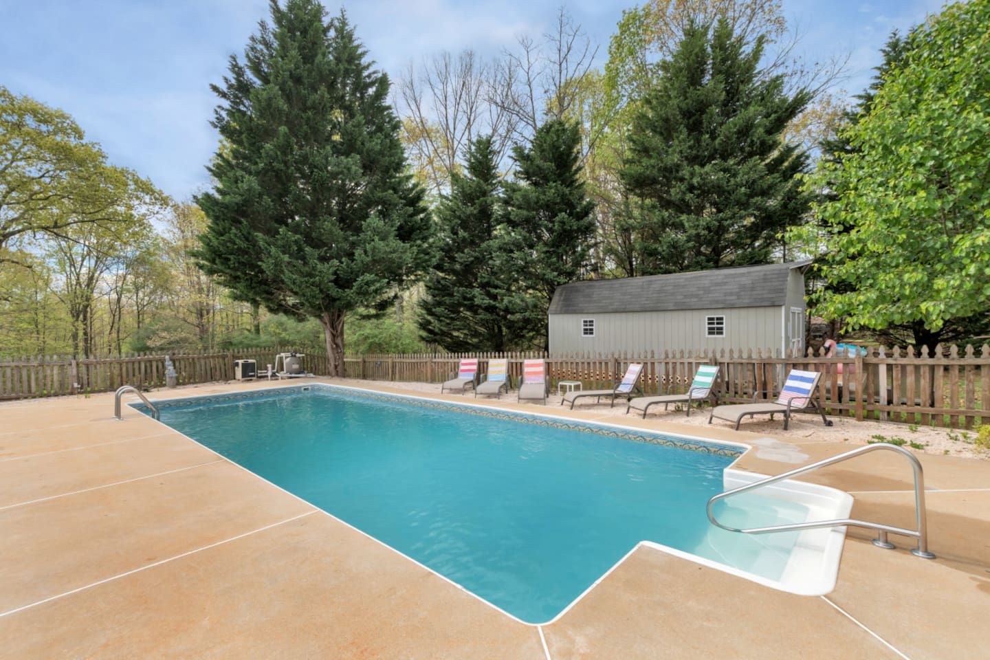 A rectangular swimming pool with a tiled deck, surrounded by a wooden fence and trees.