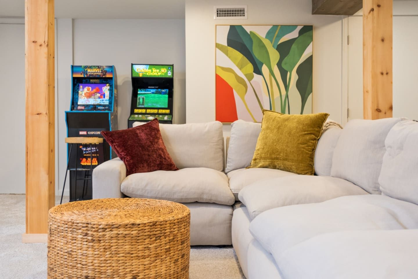 Sala de estar com sofá modular, dois consoles de jogos, um banquinho de fibra natural e um quadro abstrato colorido.