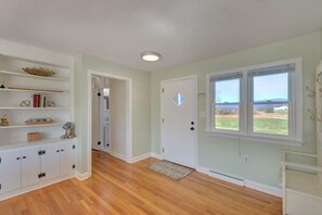 A room with wooden flooring, white walls, a door with a window, and a shelf with books and decorative items.