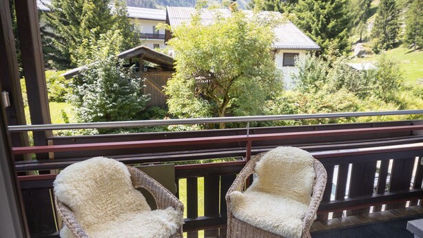 A balcony with two wicker chairs covered in white fur, overlooking a mountainous landscape with houses and trees.