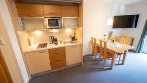 A compact kitchen with a microwave, kettle, and coffee maker, a dining area with a table and chairs, and a TV mounted on the wall.