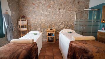 Una habitación con dos camas, una pared de piedra, un estante de madera con toallas, una mesita y una cabina de ducha de vidrio.