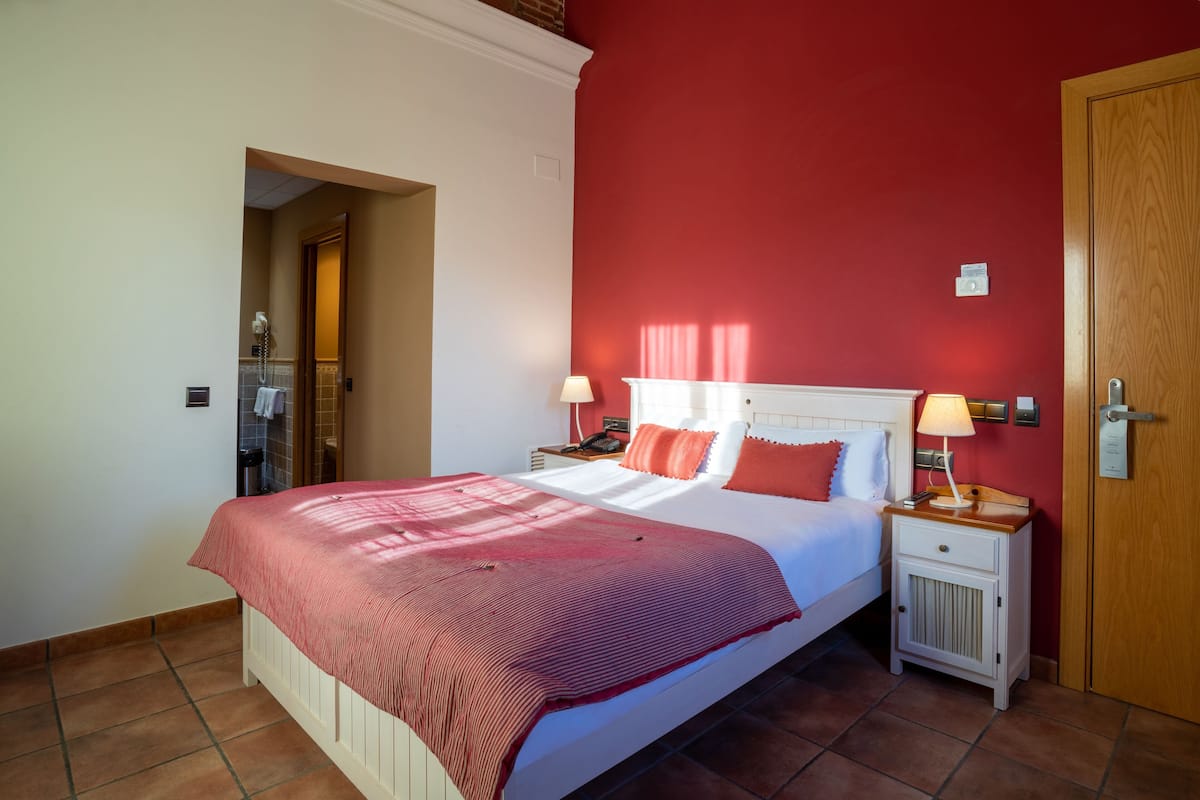 A hotel room with a bed, two bedside tables, a nightstand, a door, and a red wall.