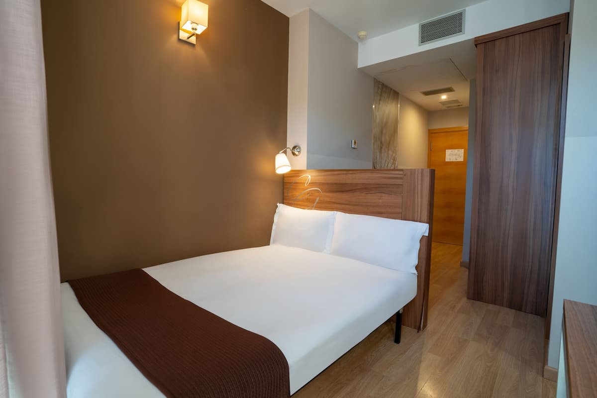 A hotel room with a bed, wooden headboard, two wall-mounted lights, a wooden door, and a wooden floor.