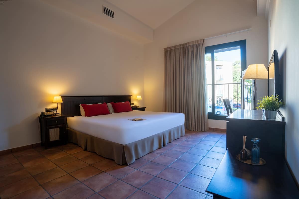 A hotel room with a large bed, two bedside lamps, a desk with a lamp, a window with curtains, and a view of the outside.