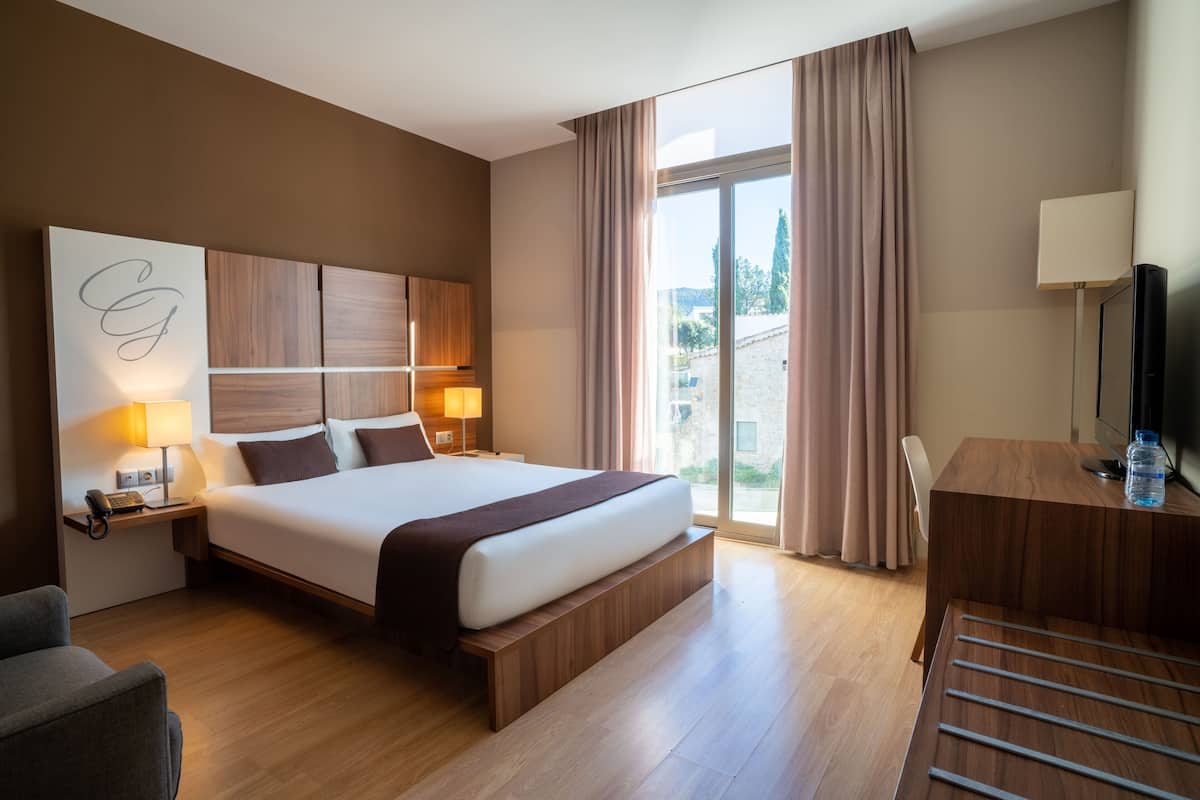 A hotel room with a large bed, a desk, a chair, a TV, and a view of the outdoors.