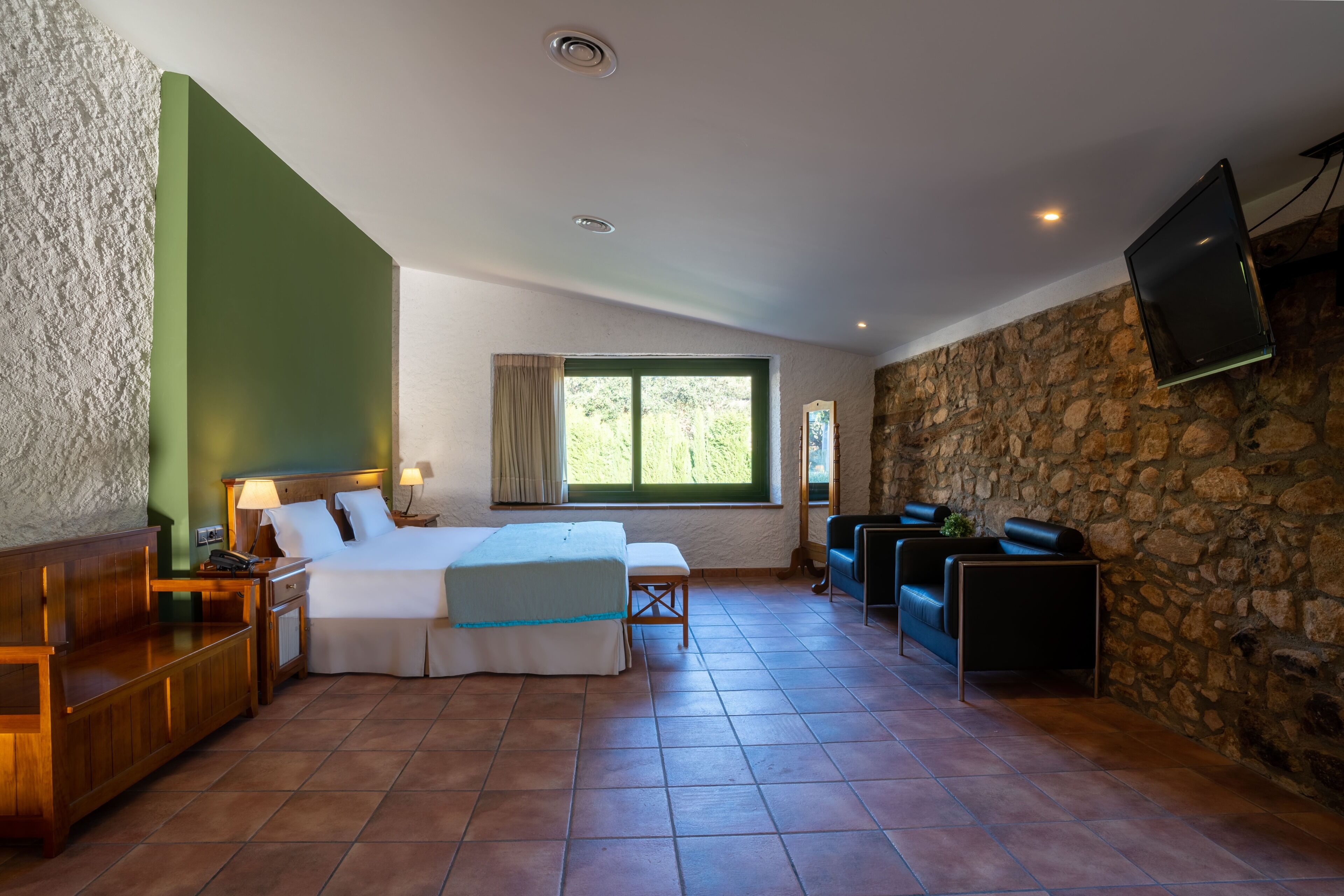 Habitación de hotel con cama, mesitas de noche, ventana, pared de piedra y televisor montado.
