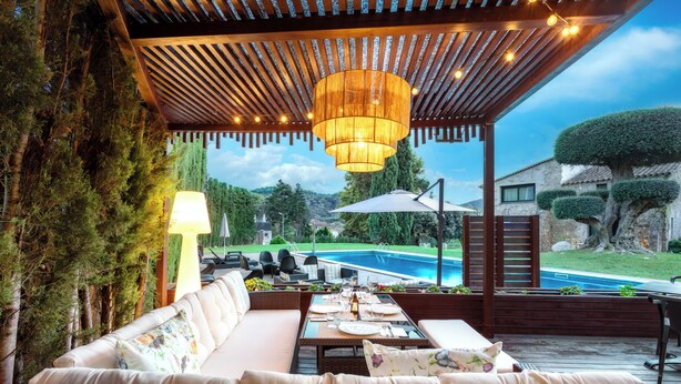 An outdoor patio with a wooden pergola, a hanging light fixture, a dining table set for two, and a view of a swimming pool. - Hotel & Spa Golden Barcelona (Vallromanes)