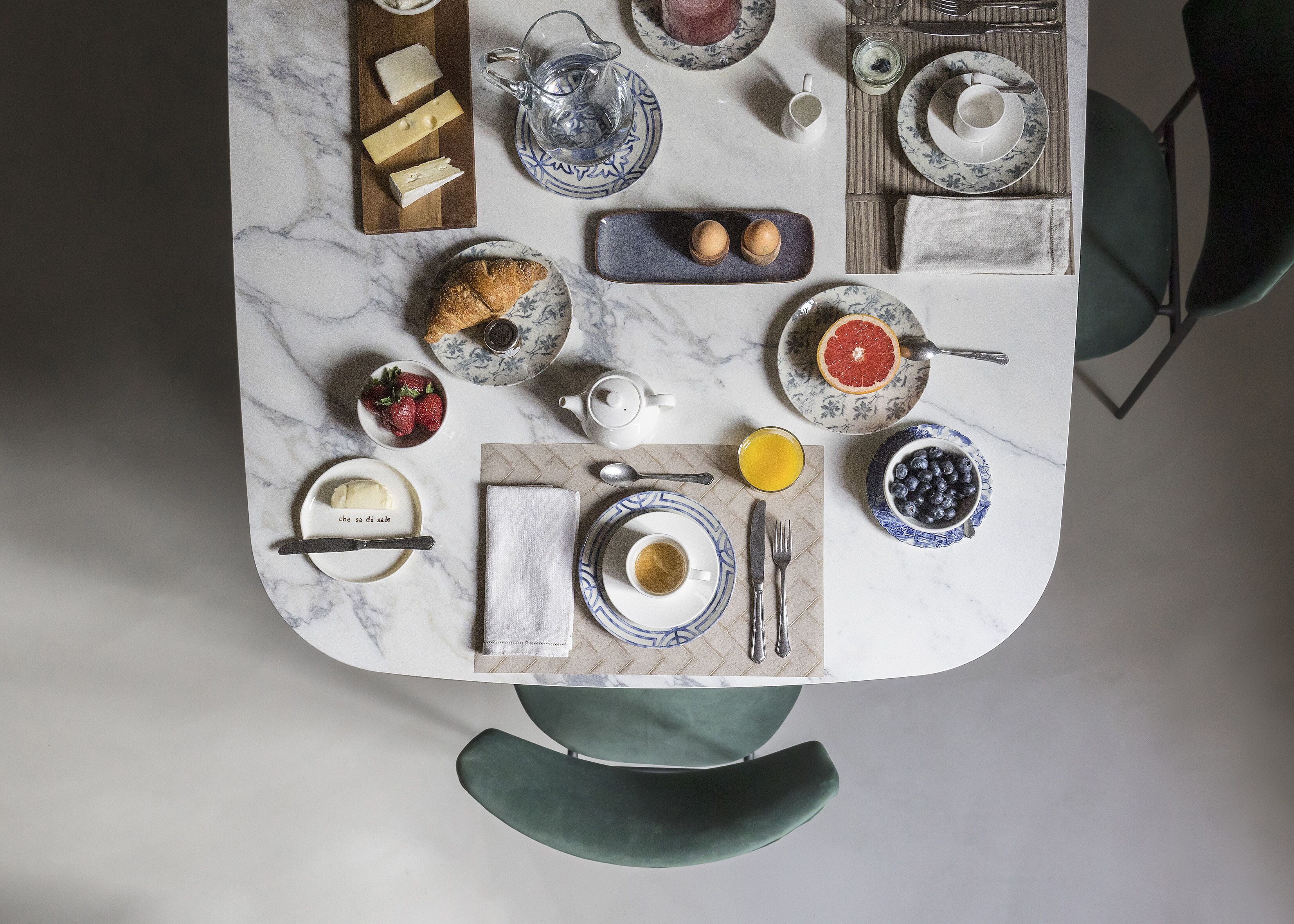Une table de petit-déjeuner dressée avec divers aliments et de la vaisselle, notamment une assiette de fromage, un bol de fraises, un croissant et une tasse de café.
