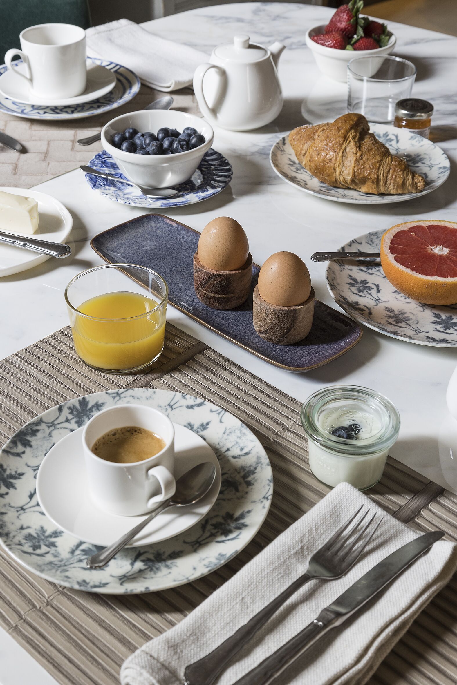 A breakfast table with a croissant, eggs, a bowl of blueberries, a glass of orange juice, and a cup of coffee.