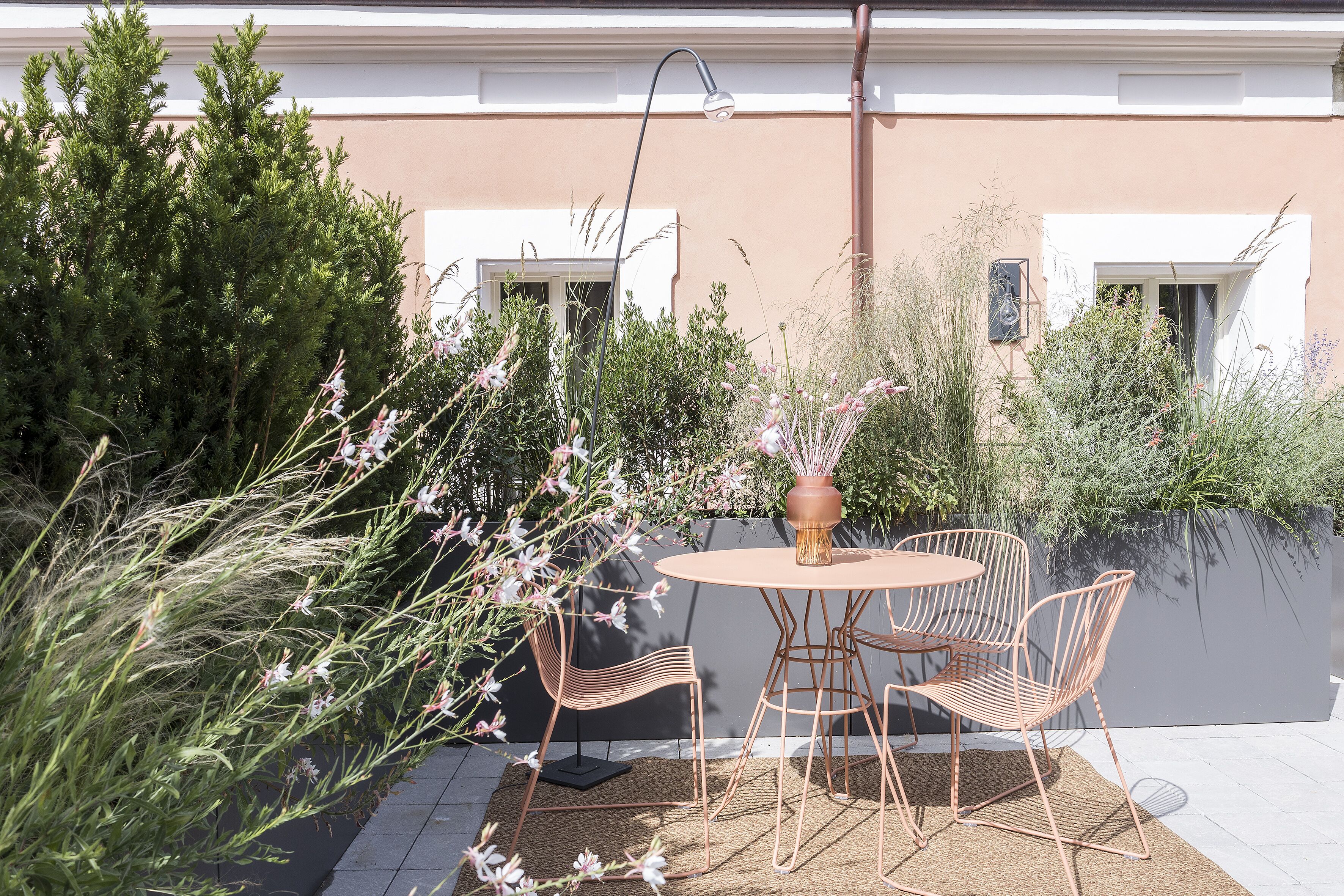 An outdoor patio with a round table and four chairs, surrounded by greenery and a building in the background.