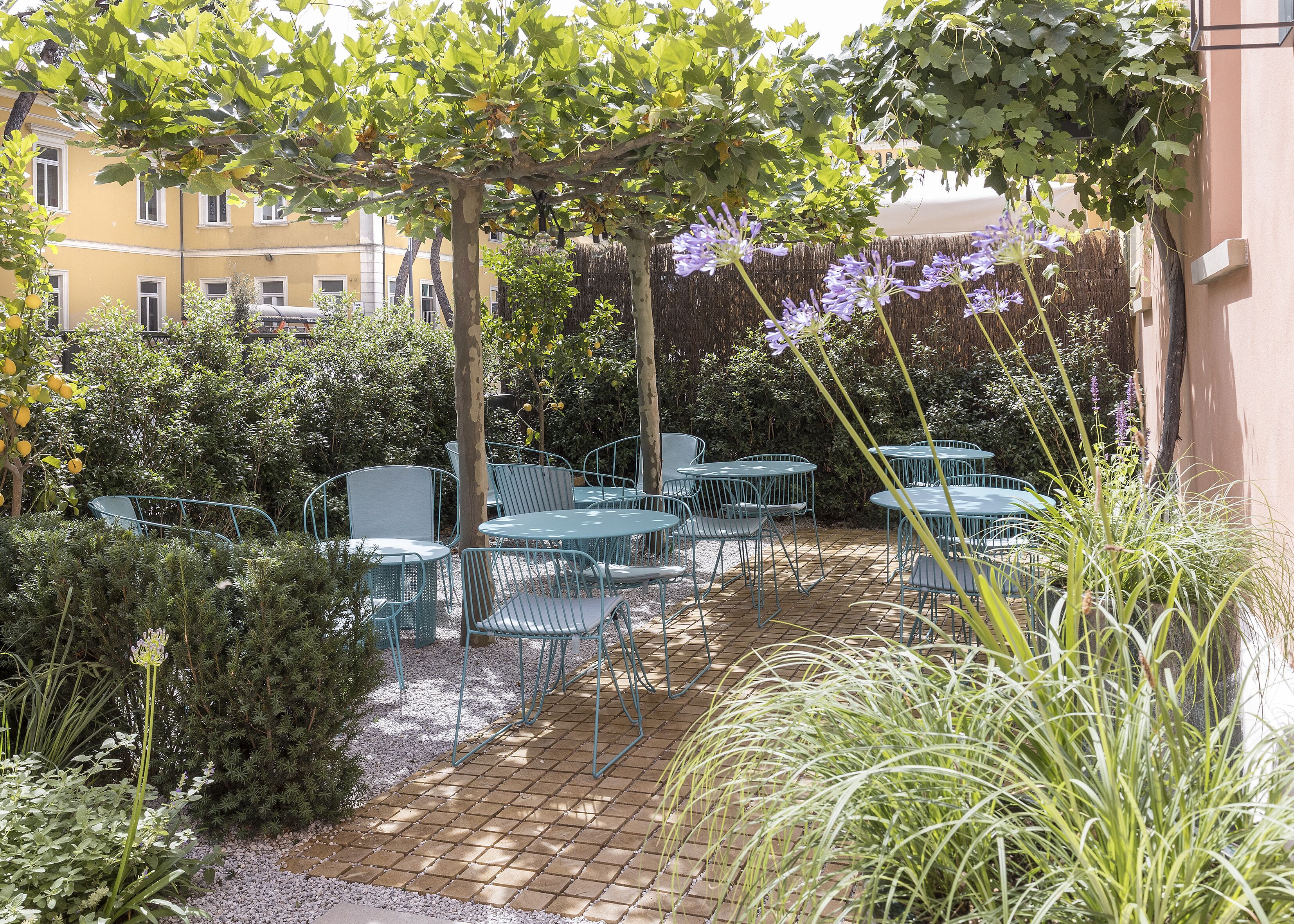 Un espace patio avec des tables et des chaises en métal, entouré de verdure et avec un bâtiment en arrière-plan.