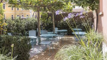 Un espace patio avec des tables et des chaises en métal, entouré de verdure et avec un bâtiment en arrière-plan.