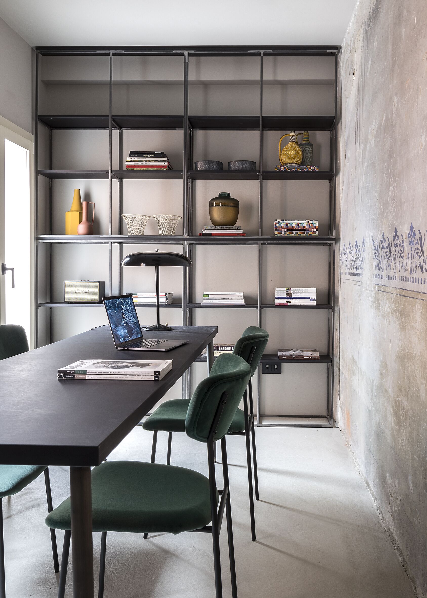 Un espacio de oficina moderno con una mesa larga, sillas verdes, una computadora portátil y una estantería llena de libros y objetos decorativos.
