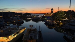 Un puerto deportivo al atardecer con varias embarcaciones amarradas, una torre destacada y agua tranquila que refleja las luces.