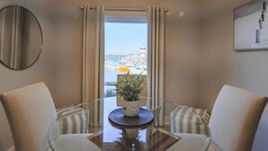 Habitación moderna con mesa de vidrio, dos sillas, una planta en maceta y vista a un faro a través de una ventana con cortinas.