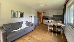 A compact living space with a grey sofa, wooden dining table, and chairs, a kitchenette with white cabinets, and a window providing natural light. - Alpenhome Apartment [6] - Apartments Hohenberger (Mallnitz)