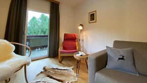 Ein Zimmer mit großem Fenster, einem roten Sessel, einem grauen Sofa und einem kleinen Tisch mit Lampe.