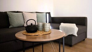 Ein runder Holztisch mit einer gewebten Tischmatte, einer schwarzen Kanne und zwei grauen Tassen auf einem grauen Sofa mit weißer Tagesdecke.