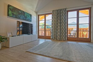 A living room with a flat-screen TV, a mounted artwork, a large window with a view of a mountain, and a sliding glass door. - Ferienwohnung- 3 Personen, 68qm, Wohnküche, 1 Schlafzimmer, Wlan (Marquartstein)