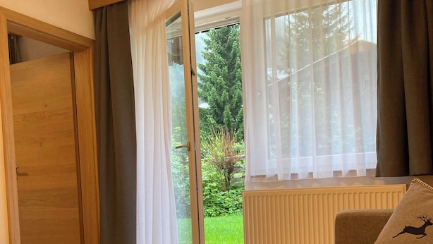 A room with a wooden door, a window with sheer curtains, a view of greenery outside, and a sofa with a cushion featuring a deer silhouette.