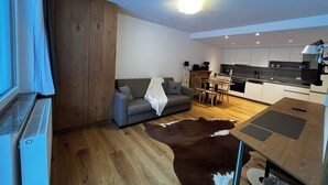 A modern living room with a grey sofa, wooden flooring, a cowhide rug, and a kitchen area with white cabinets and a dining table.