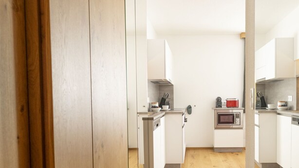 Un estrecho pasillo con piso de madera que conduce a una zona de cocina con armarios blancos y un microondas.