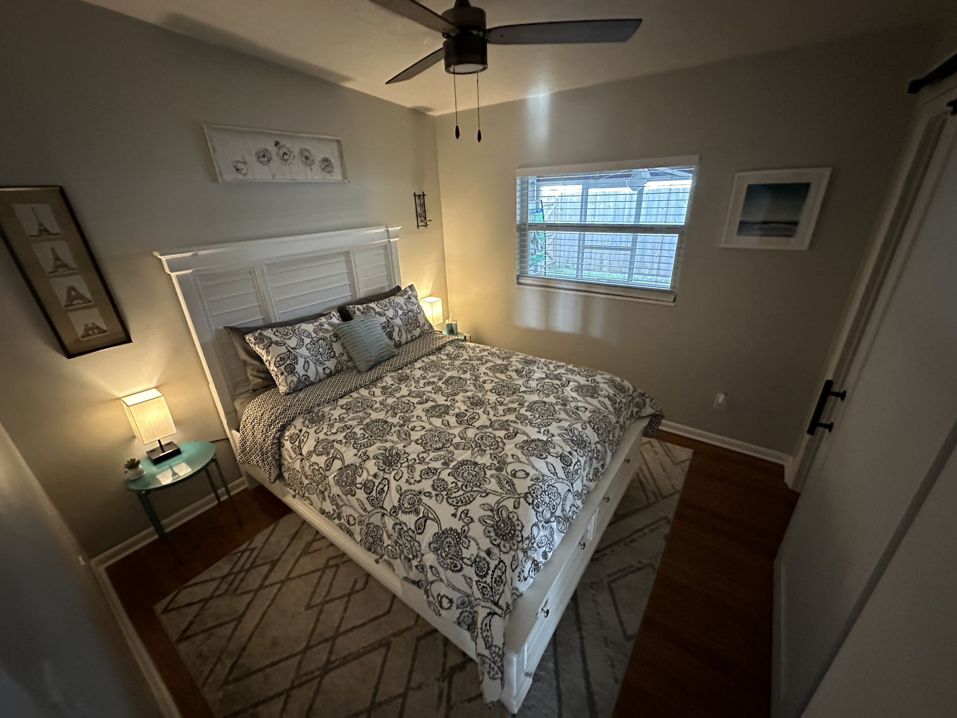 A bedroom with a bed, a ceiling fan, a window, and a nightstand with a lamp.