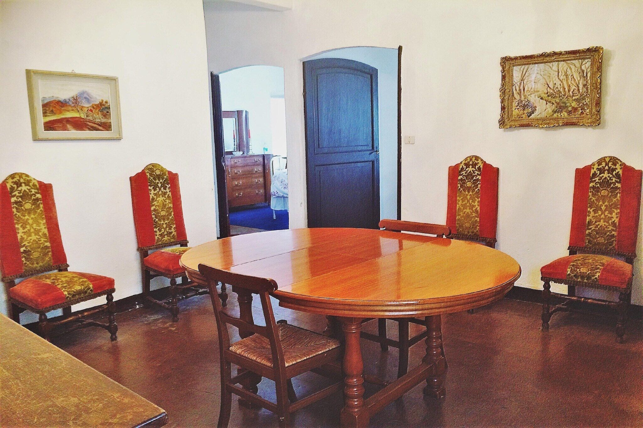 A dining room with a round wooden table and four chairs with red cushions. There are two framed pictures on the walls.